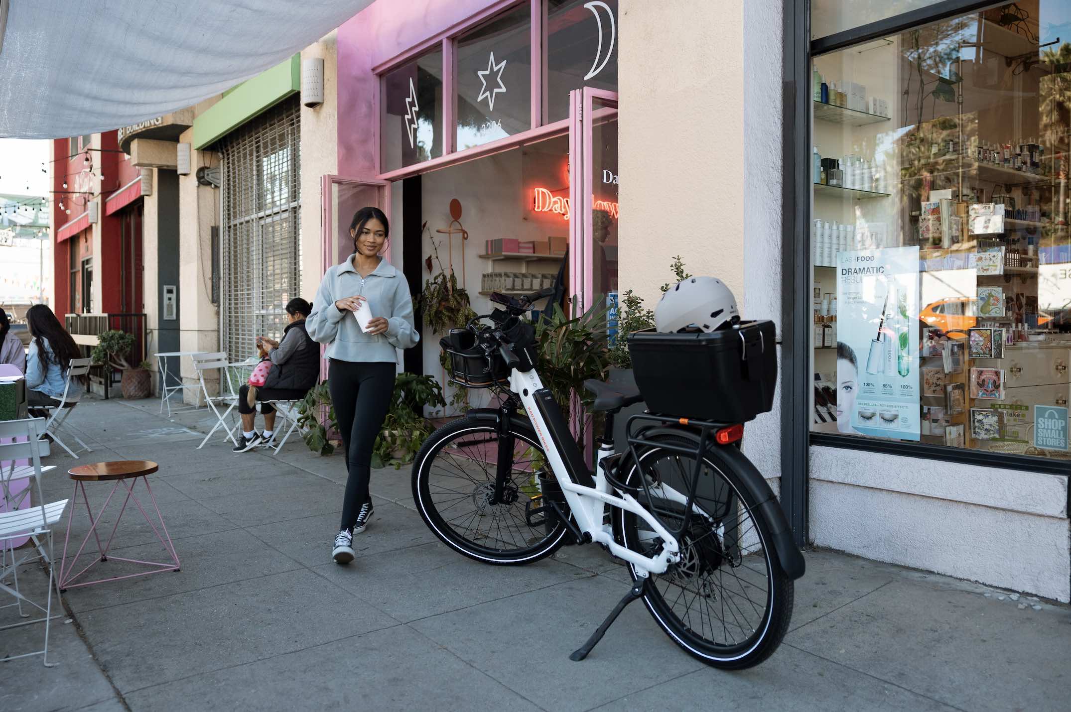 A woman leaves a cafe with a cold drink and walks towards her white Radster Road ebike.